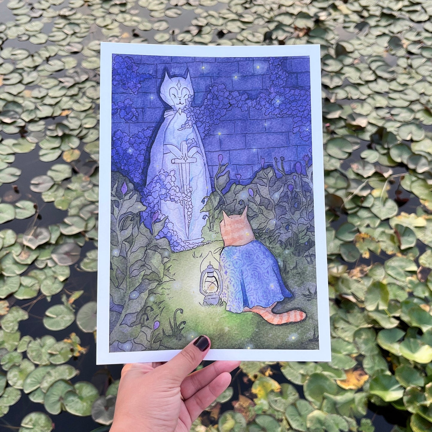 A hand holds a square art print featuring a whimsical illustration of a white cat in a blue dress standing in a garden at night, with a small orange cat sitting beside it. The background is a real outdoor pond covered with green lily pads.