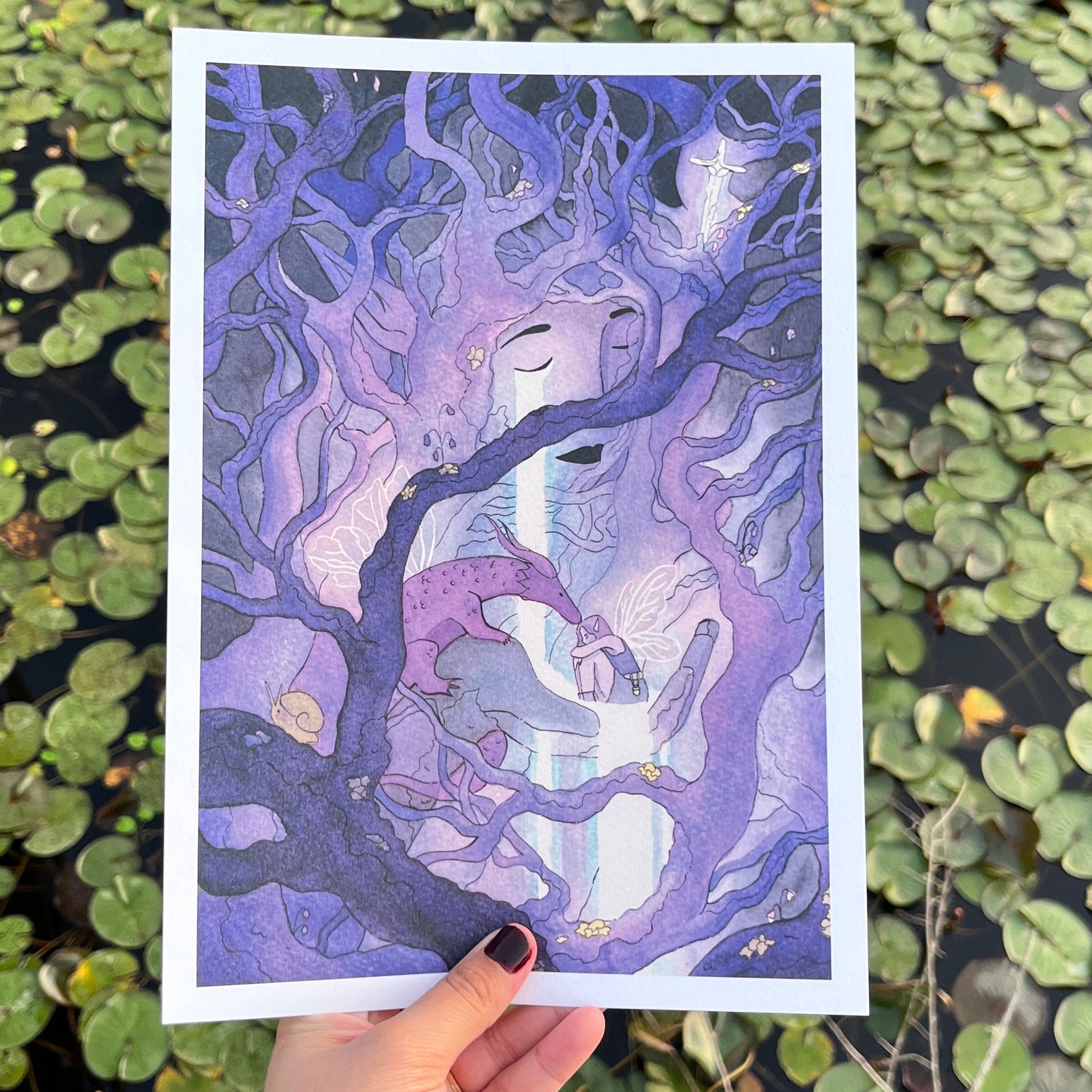 A hand holds a square art print featuring a purple and blue illustration of a tree with a face, set against a background of green lily pads on water.