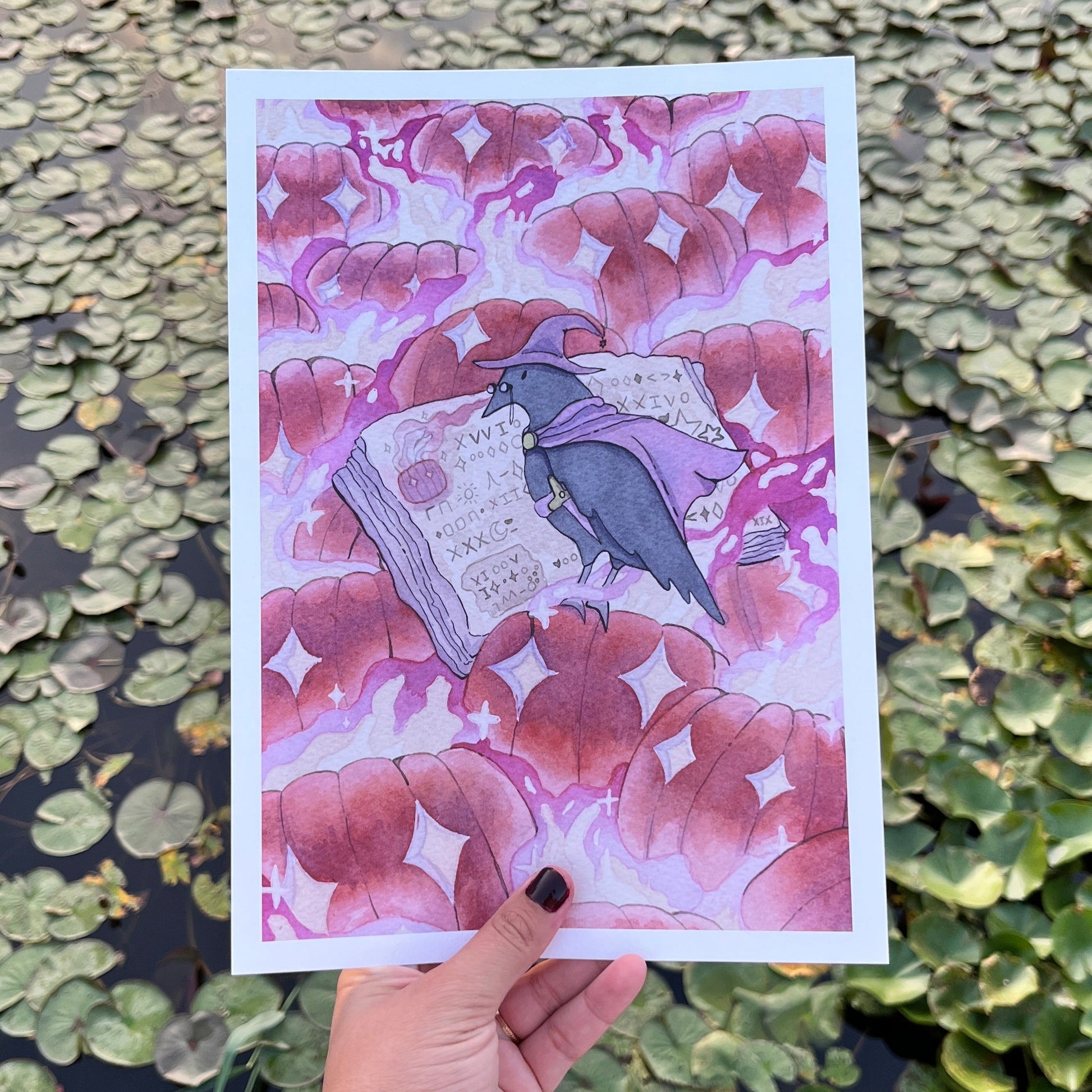 A hand holds a square art print featuring a purple and pink illustration of a raven perched on an open book, set against a background of green lily pads on water.