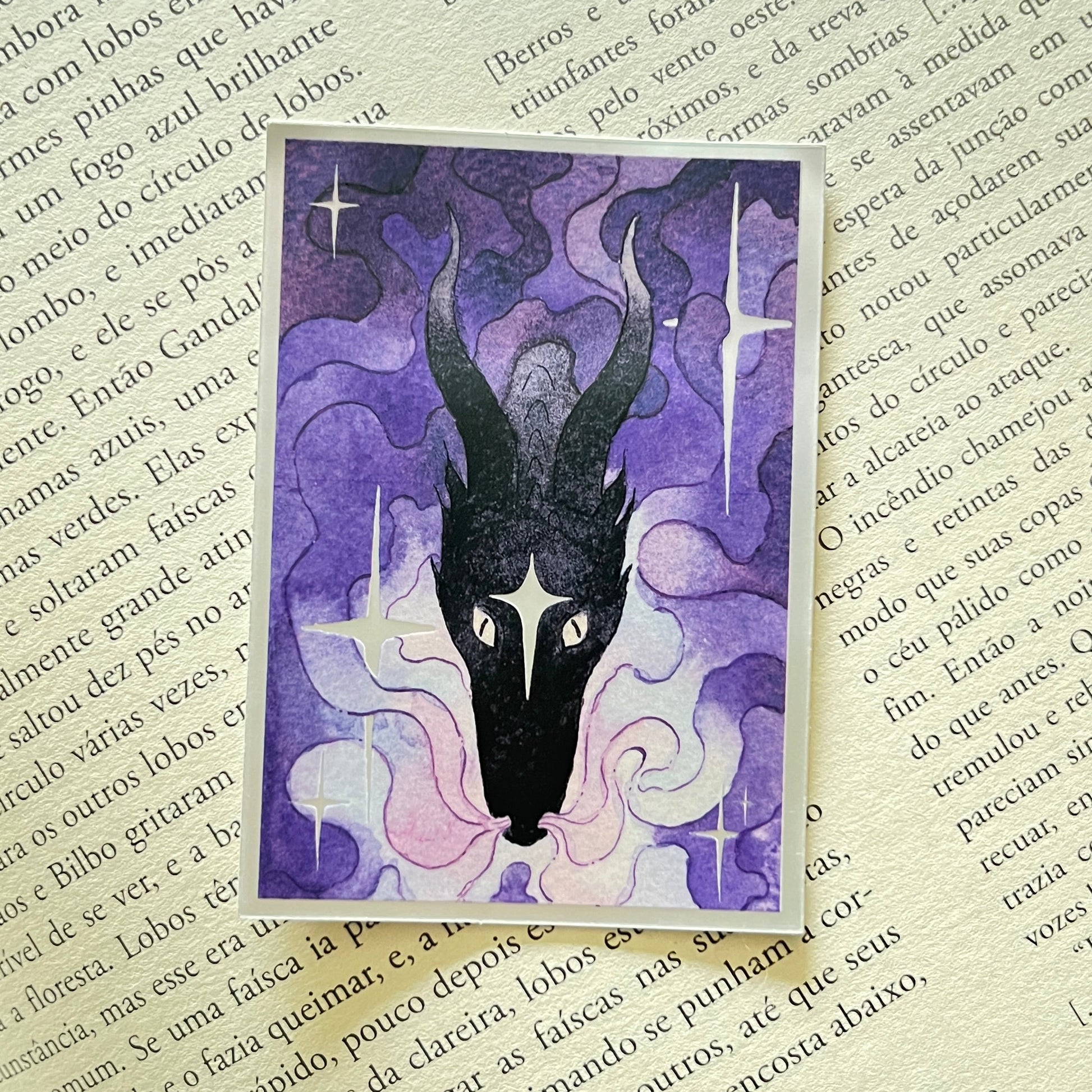 A rectangular art print featuring a stylized black dragon head with long horns on a purple and pink background, placed on top of an open book with printed text.